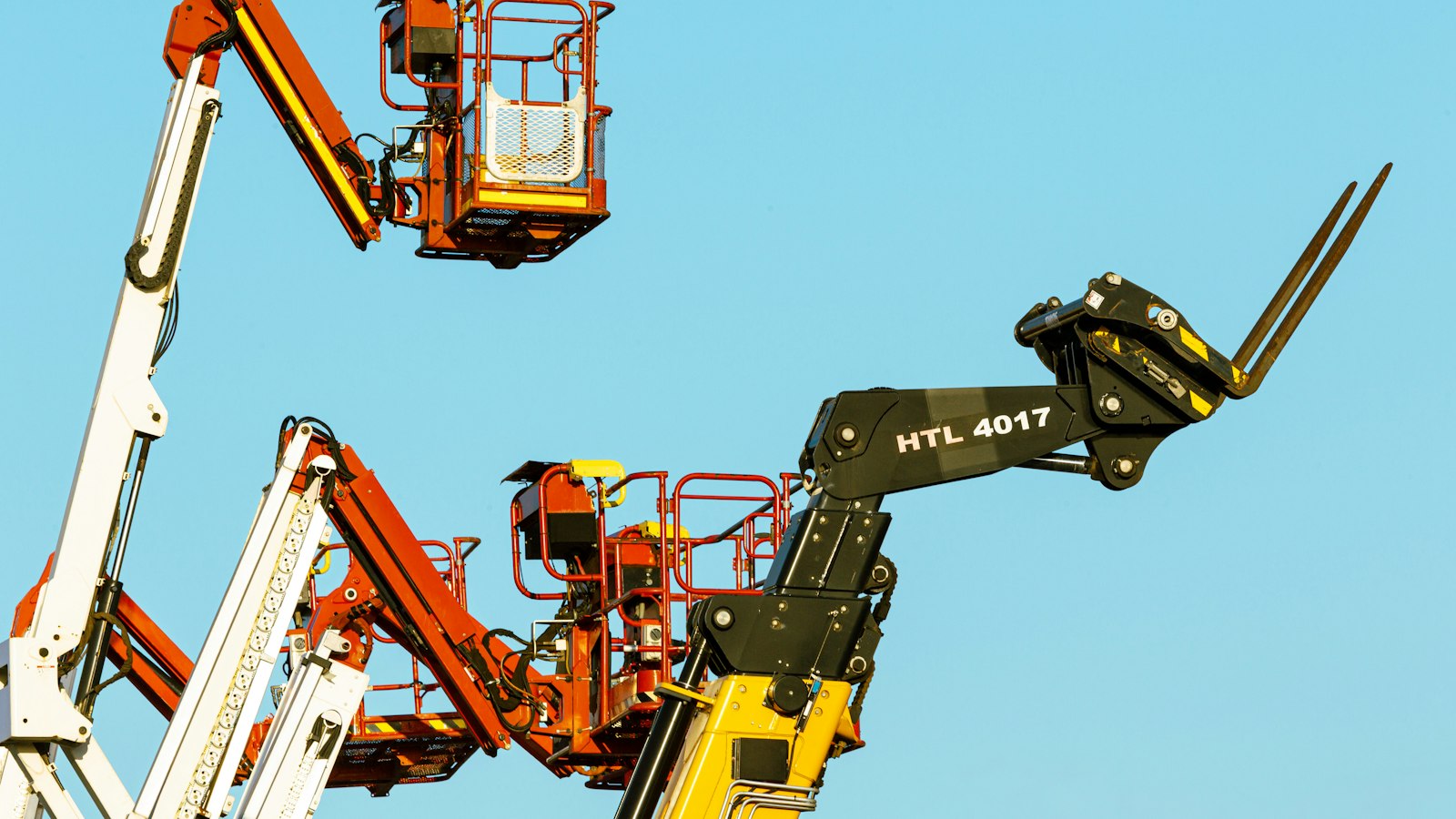 Two red aerial work platforms and a black telehandler raised against a clear blue sky, the kind of rental fleet equipment that sits unreturned on stalled jobsites