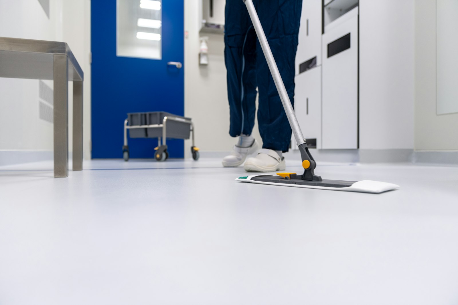 A facility services worker in blue scrubs running a flat mop over a white floor in a clinical hallway, the kind of recurring contract cleaning that keeps getting done while the monthly invoices age