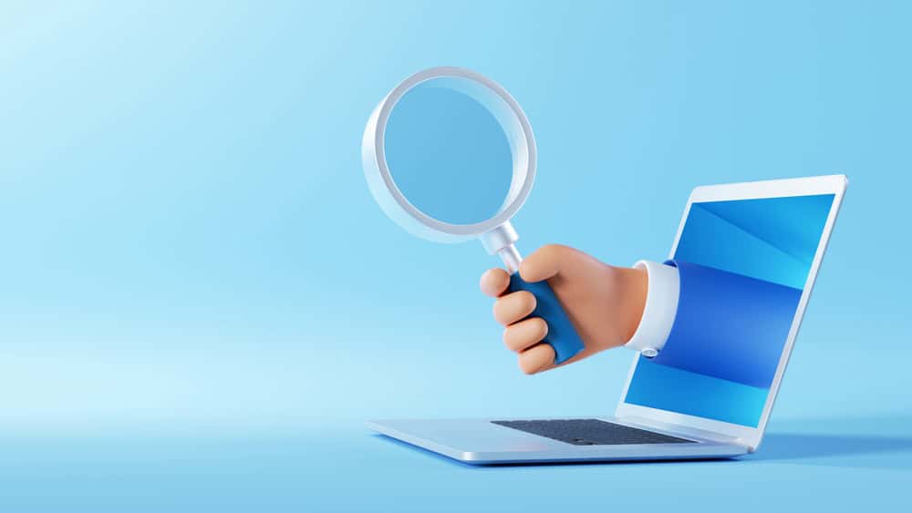 Hand holding a magnifying glass out of a laptop screen, representing skip tracing and debtor investigation