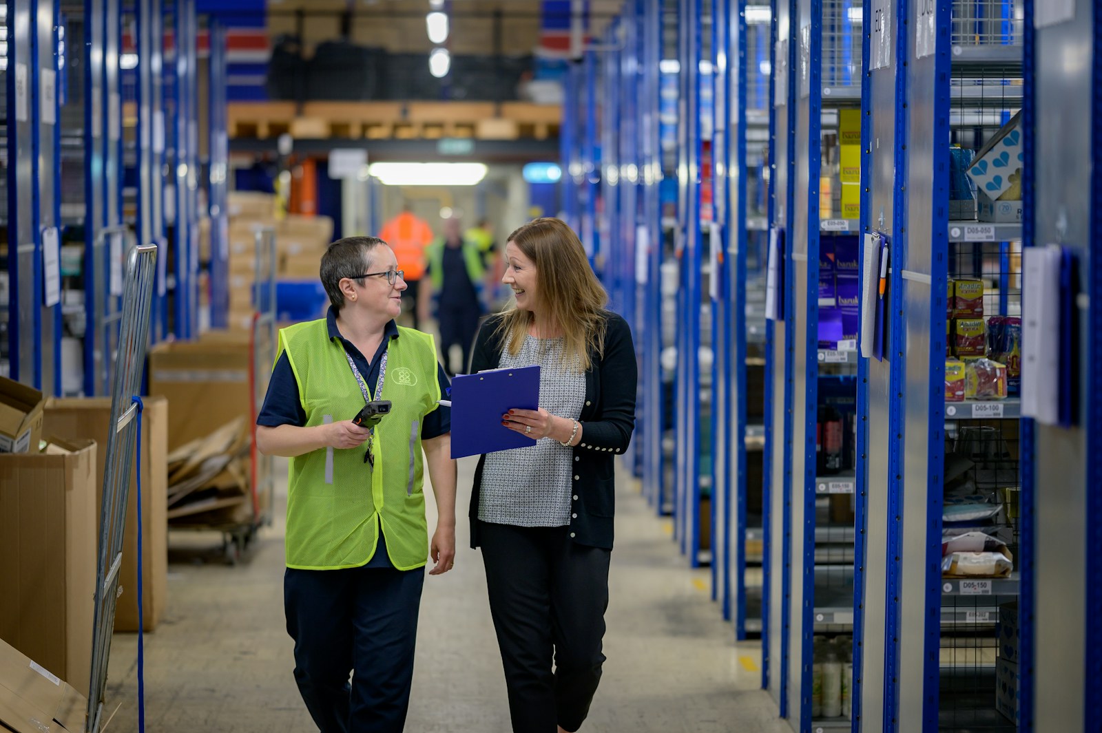 A warehouse worker in a high visibility vest and a colleague holding a clipboard walking a stocked warehouse aisle, the kind of client site where staffing firms place temporary workers